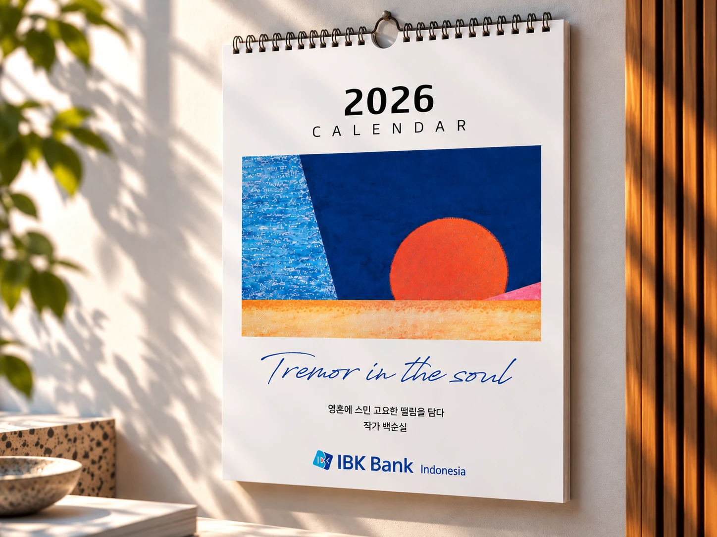 IBK Bank Indonesia 2026 wall calendar hanging on a sunlit wall, the cover artwork a sun over water.